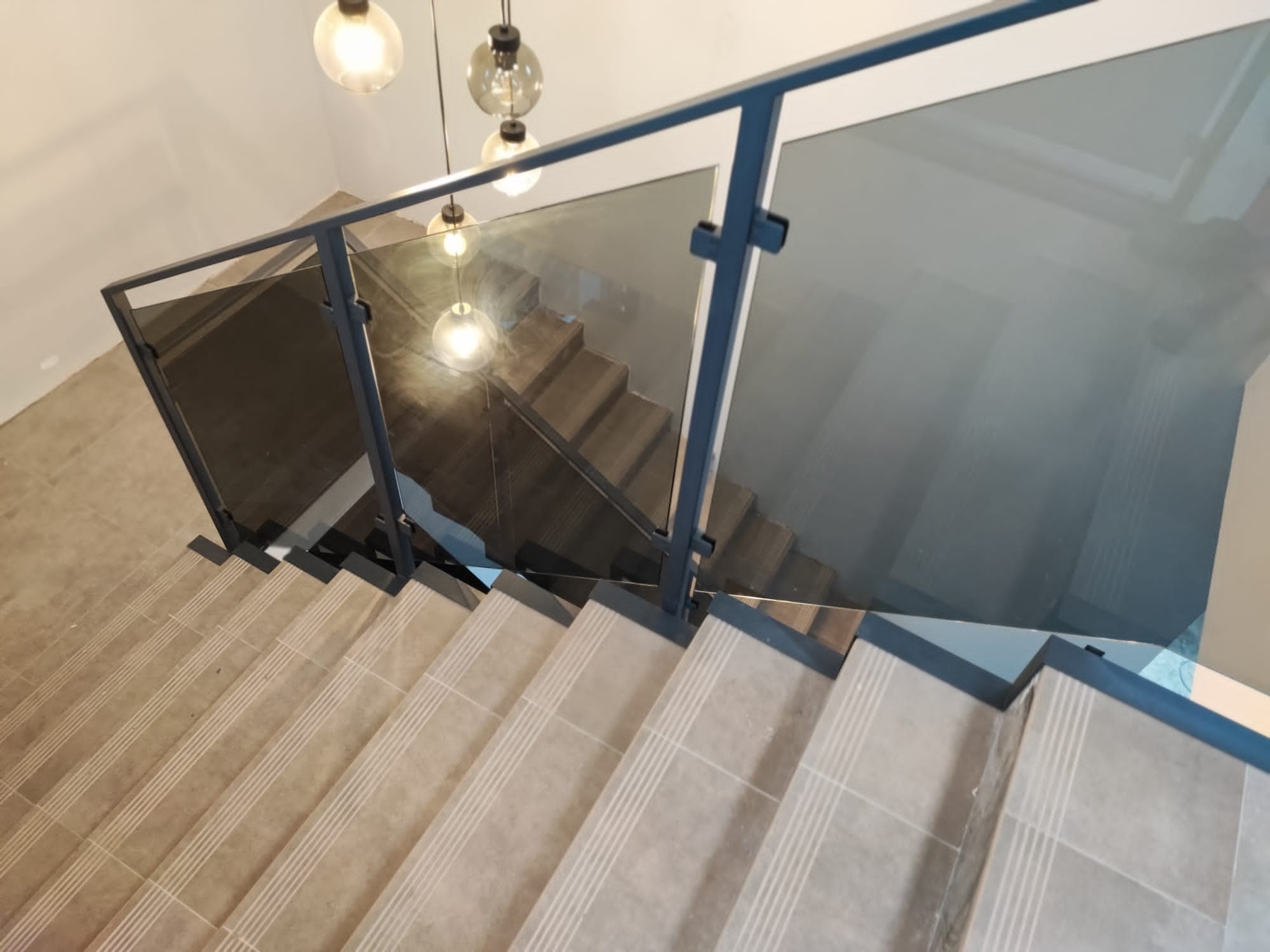 Szklana balustrada z ciemną ramą, widok z góry na schody z płytkami w odcieniach beżu. Nad schodami wiszą nowoczesne lampy.