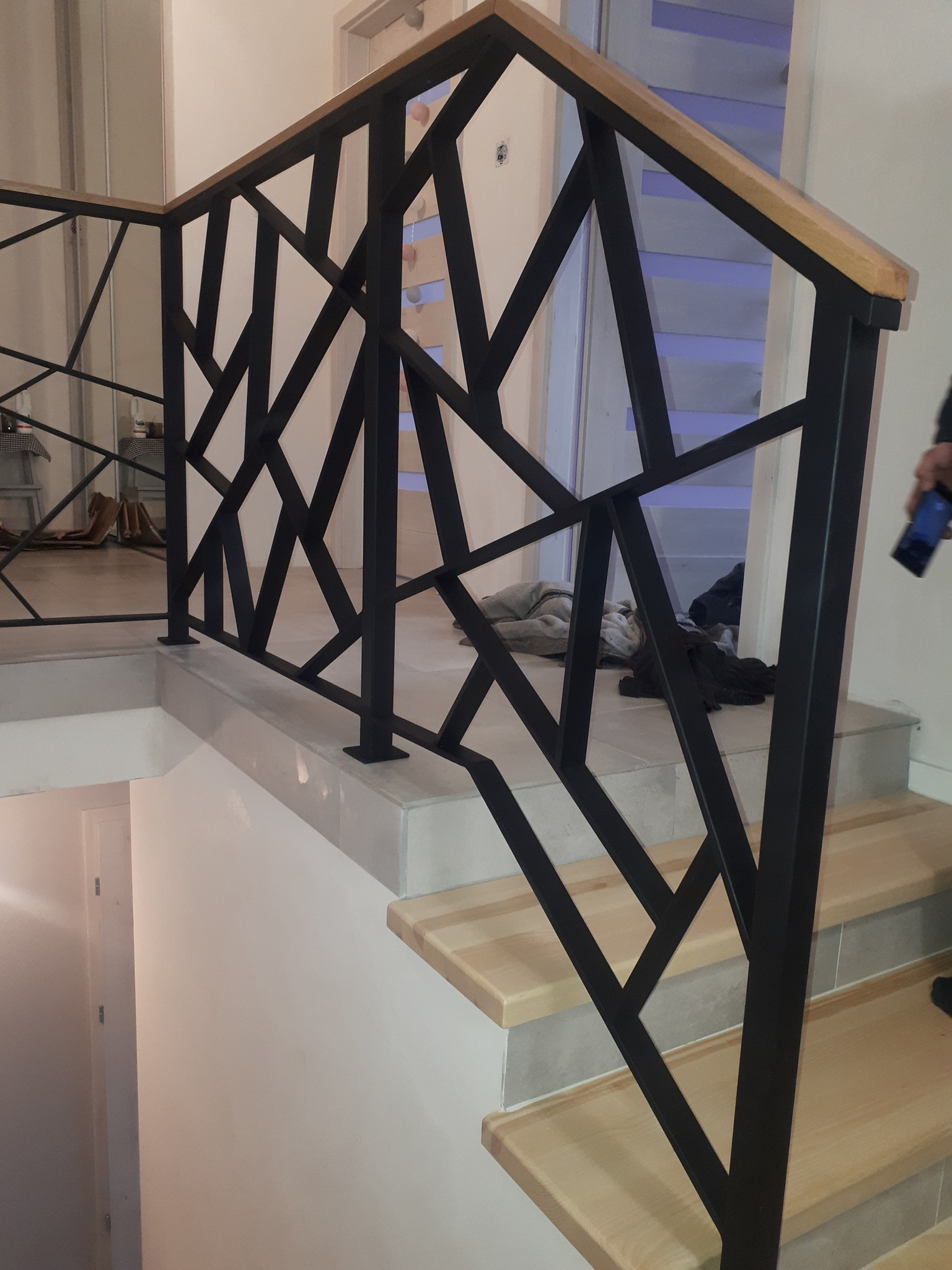 Nowoczesna balustrada schodowa z czarnego metalu o geometrycznym wzorze, wykończona drewnianym poręczą, zamontowana na jasnych schodach wewnątrz budynku.