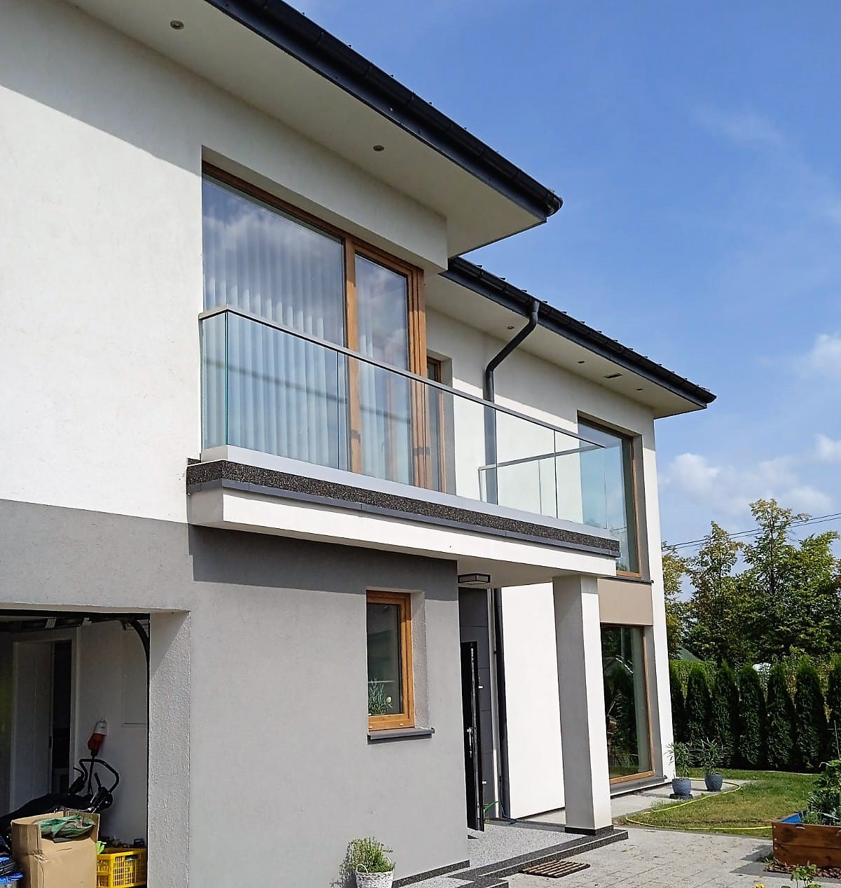 Nowoczesny dom z balkonem zabezpieczonym szklaną balustradą, widok z zewnątrz, błękitne niebo w tle. Minimalistyczny design i jasna kolorystyka budynku.