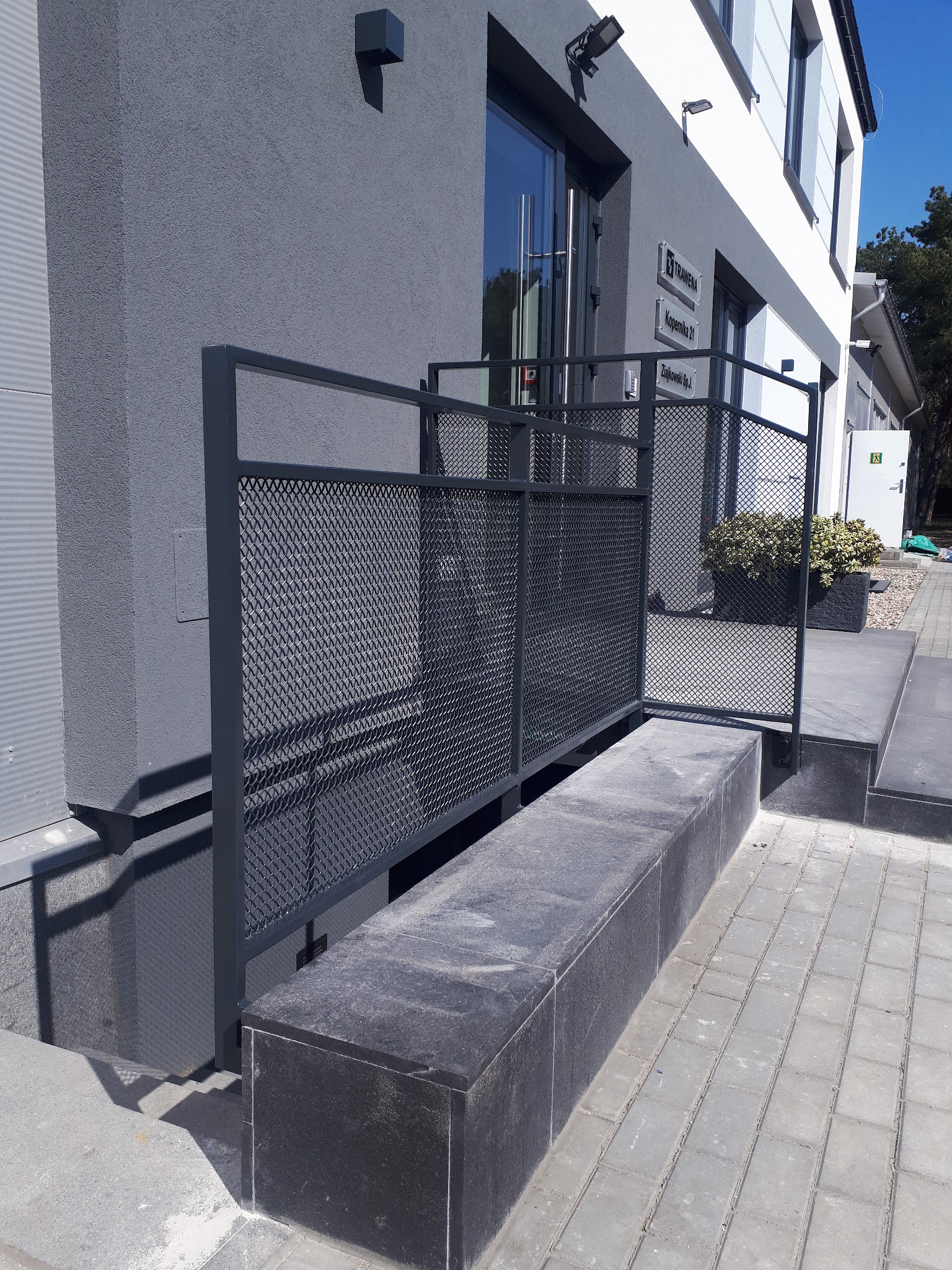 Nowoczesna balustrada metalowa z siatką, zabezpieczająca zejście do piwnicy przy budynku biurowym. Szara kolorystyka, minimalistyczny design.