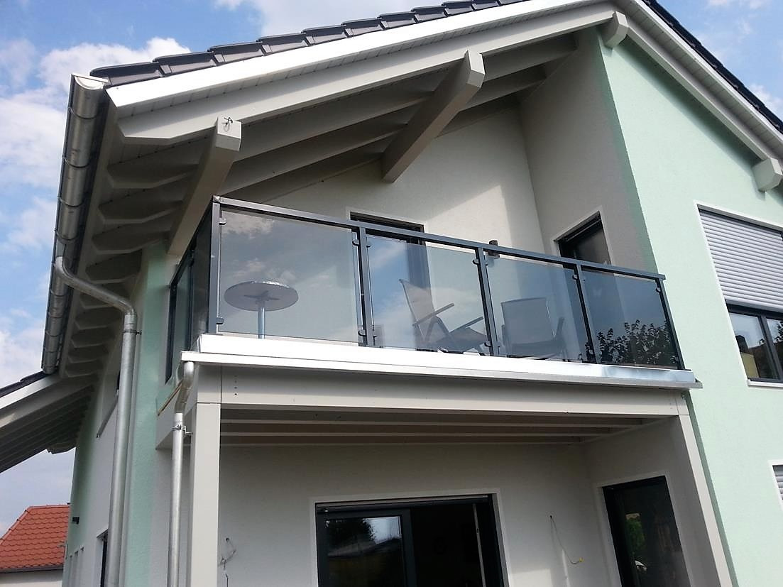 Szklana balustrada na balkonie domu z jasną elewacją i szarym dachem. Widoczne krzesła i stolik na balkonie. Ujęcie z dołu, błękitne niebo w tle.