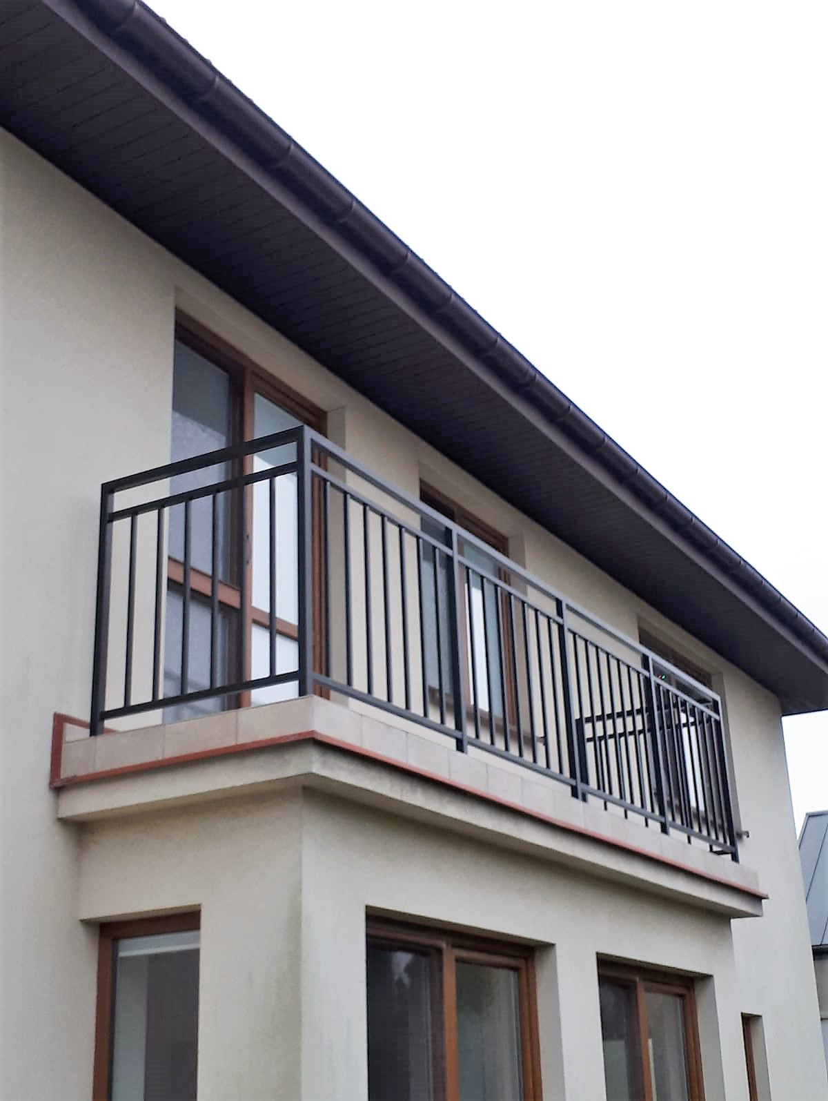 Czarna, metalowa balustrada balkonowa z pionowymi prętami, zamontowana na jasnym budynku z brązowymi oknami i ciemnym dachem. Ujęcie z dołu, podkreślające solidność konstrukcji.