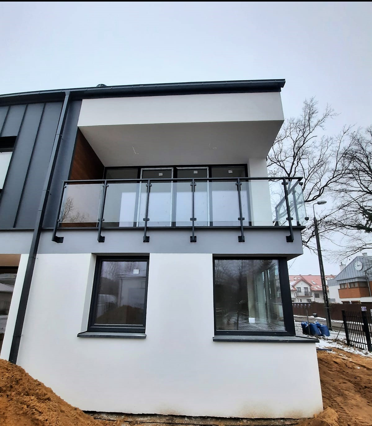 Nowoczesny budynek z balkonem zabezpieczonym szklaną balustradą z czarnymi elementami mocującymi. Widok z dołu, na pierwszym planie ziemia.