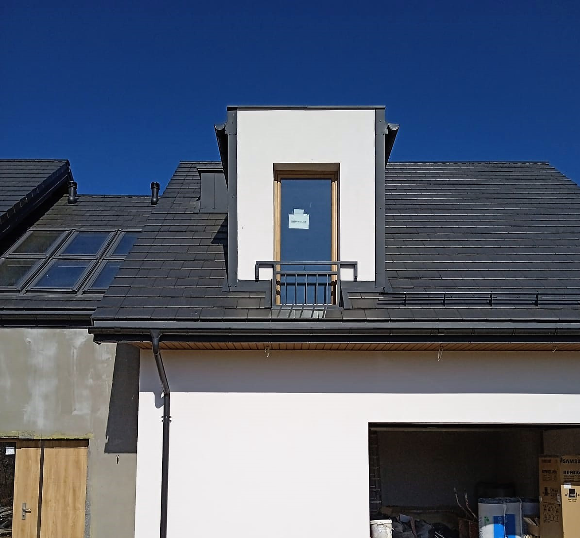 Nowoczesny dom z ciemną dachówką i oknem mansardowym z metalową balustradą. Widoczne elementy wykończeniowe i fragment garażu. Błękitne niebo w tle.