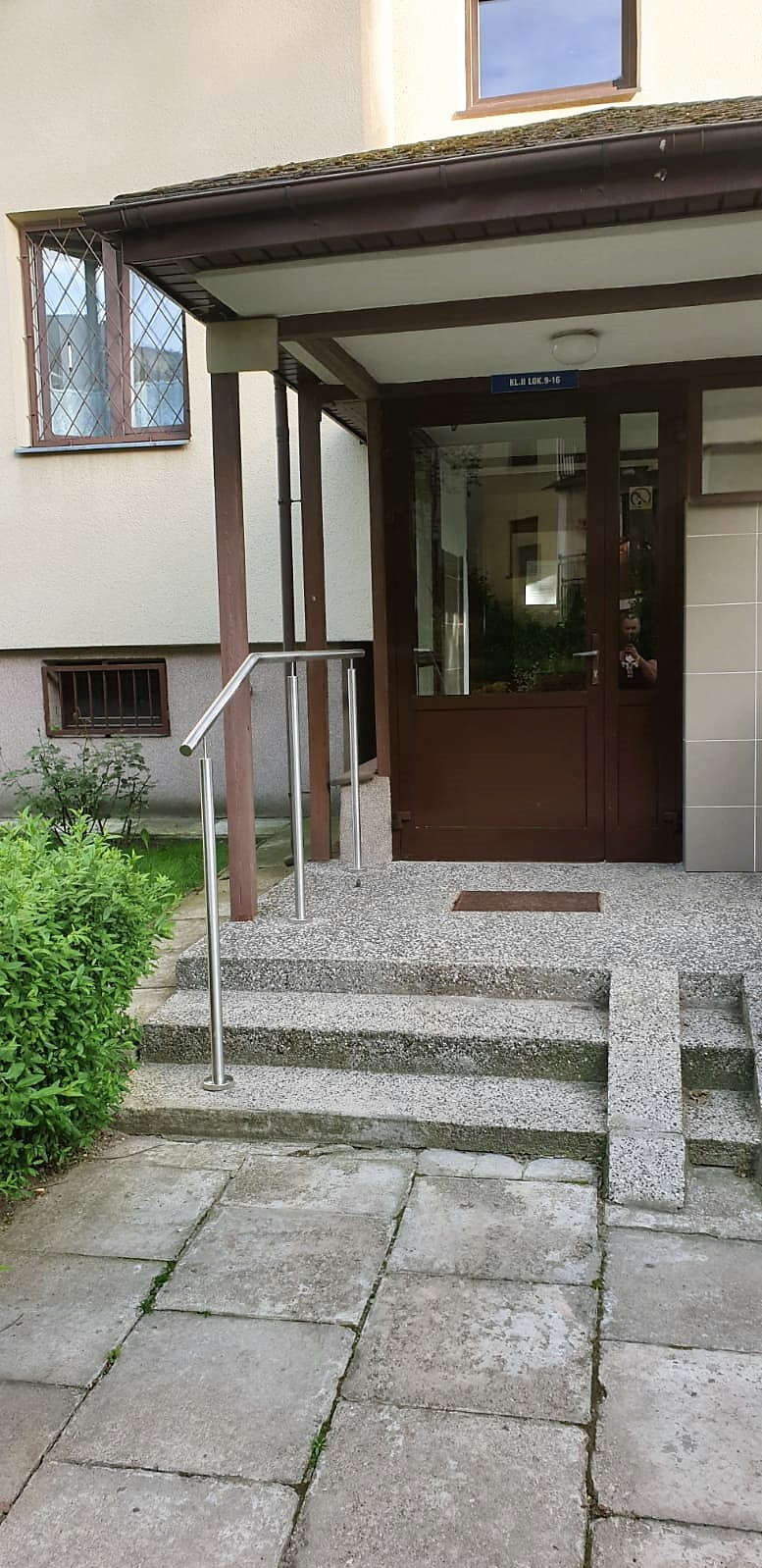 Wejście do budynku z brązowymi drzwiami i metalową balustradą przy schodach. Widoczne płytki chodnikowe i zieleń. Nad drzwiami tabliczka z numerem lokalu.