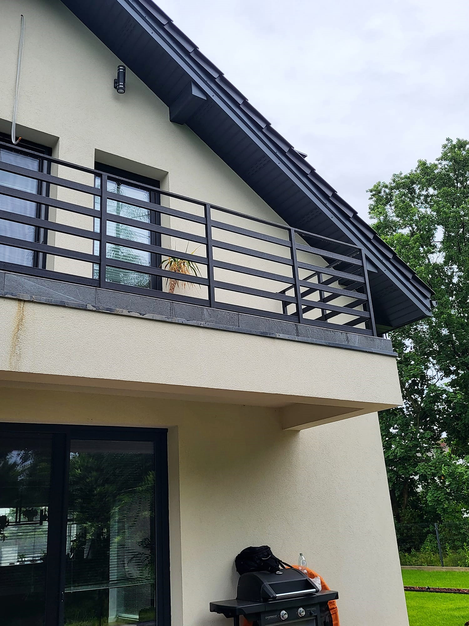 Nowoczesna balustrada balkonowa z czarnych, poziomych profili na tle jasnej elewacji domu z ciemnym dachem i zielenią. Widok z dołu, podkreślający minimalistyczny design.