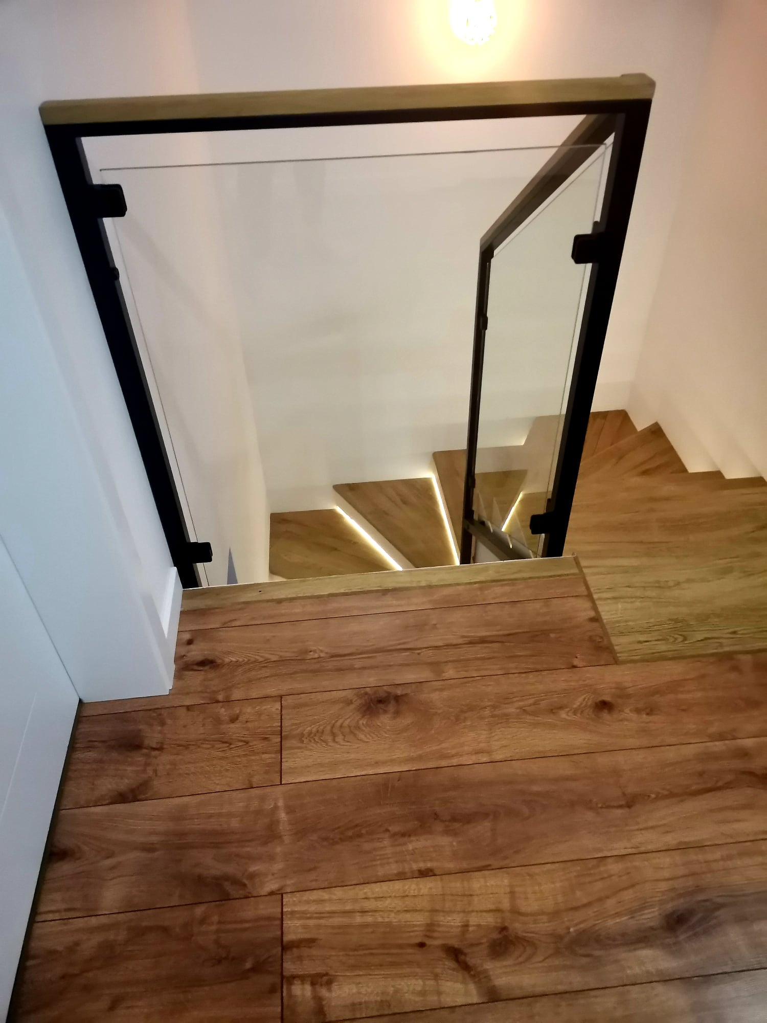 Nowoczesne schody drewniane z podświetleniem LED i szklaną balustradą z czarnymi elementami, widok z góry na drewnianą podłogę.