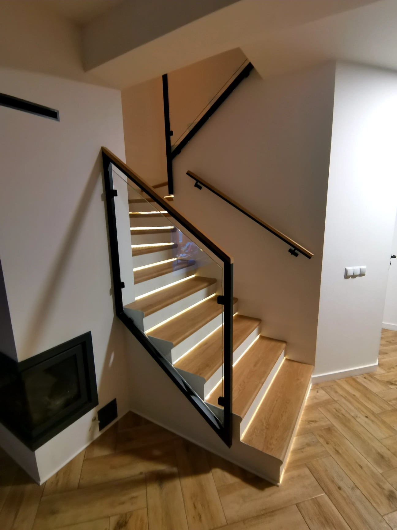 Nowoczesne schody drewniane z podświetleniem LED i balustradą ze szkła hartowanego, czarne metalowe elementy konstrukcyjne, drewniana podłoga w jodełkę.
