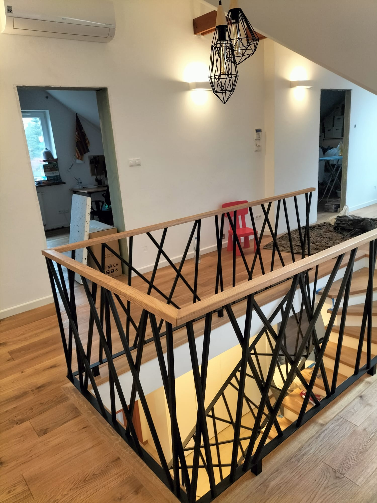Nowoczesna balustrada schodowa z czarnego metalu i drewna, z geometrycznym wzorem, na tle jasnego wnętrza z drewnianą podłogą i widocznymi schodami.