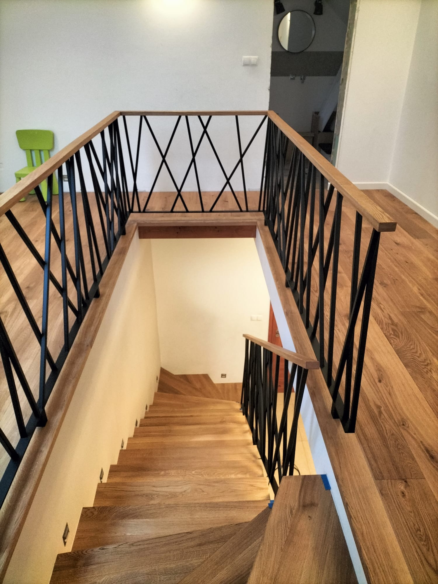 Drewniane schody z czarną, metalową balustradą o geometrycznym wzorze, widziane z góry. Jasne drewno stopni i poręczy kontrastuje z ciemną balustradą.