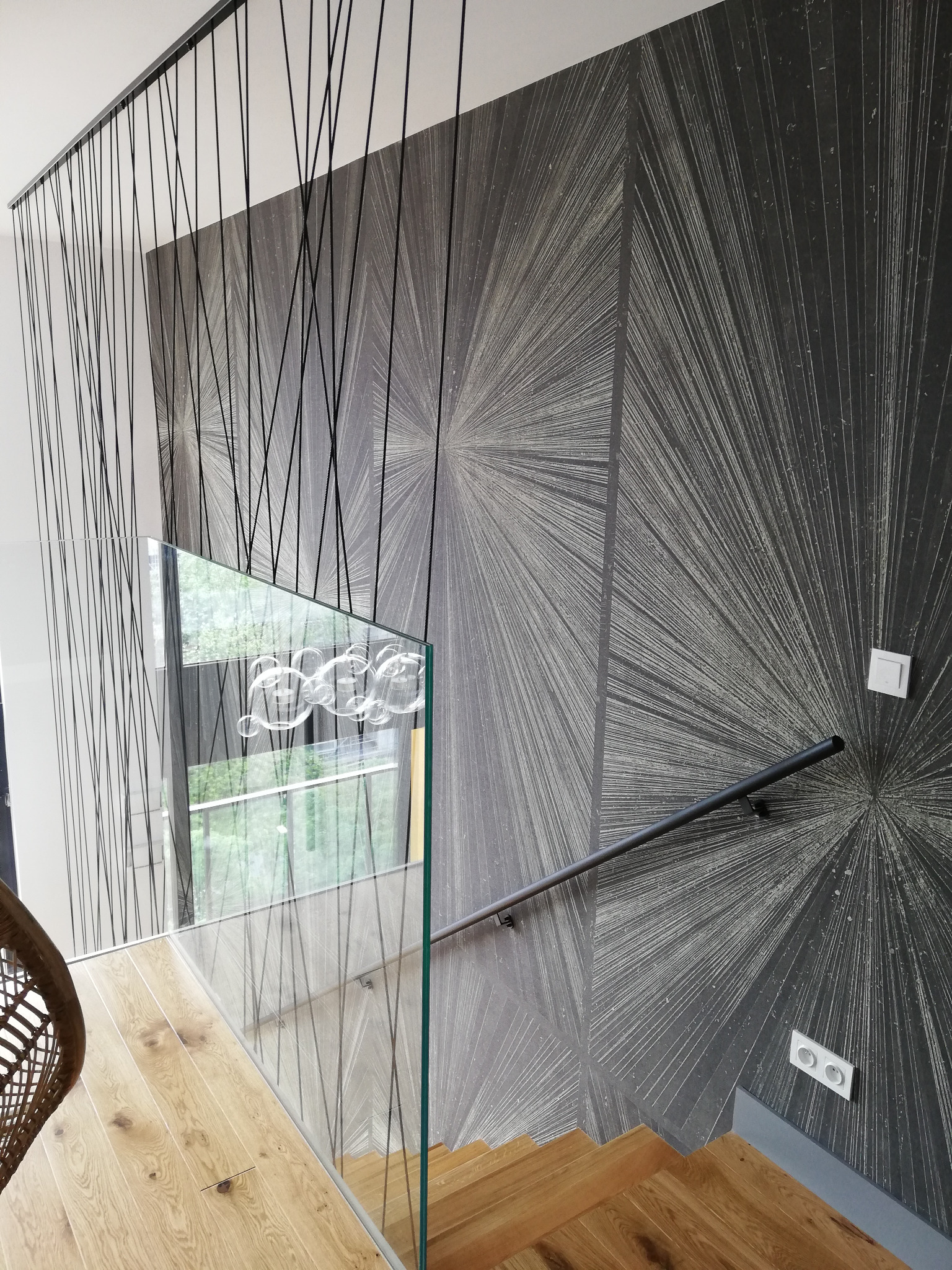 Szklana balustrada schodowa z drewnianymi stopniami i czarną poręczą na tle ściany z geometrycznym wzorem. Wiszące czarne sznurki dodają nowoczesnego charakteru.