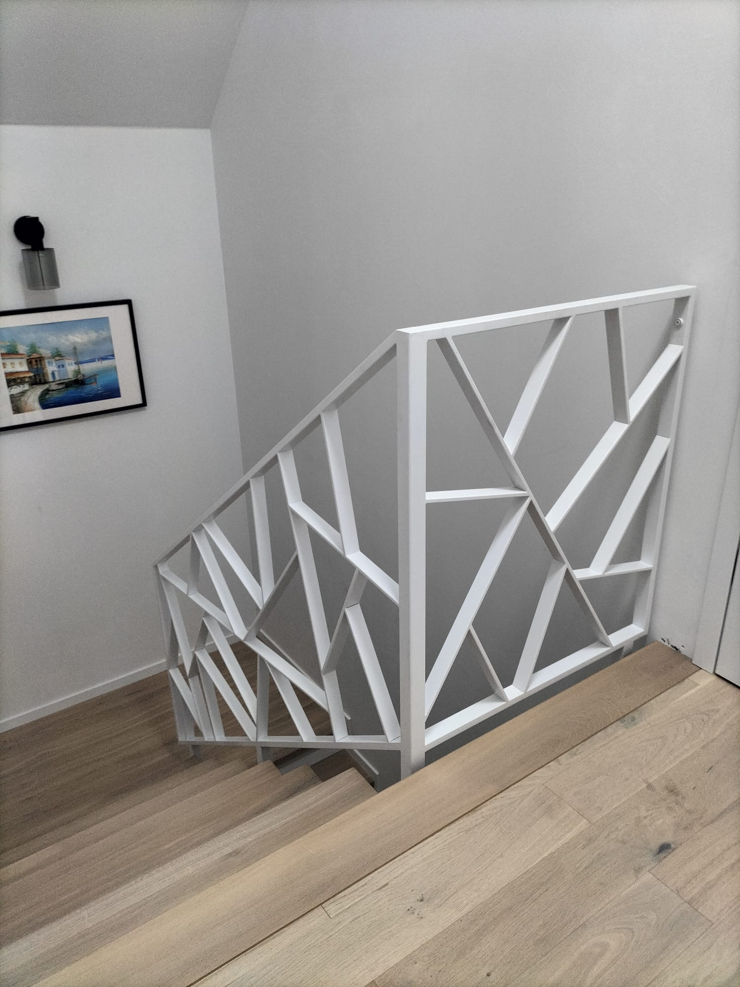 Biała balustrada schodowa o geometrycznym wzorze, widok z góry na drewniane schody. Na ścianie obraz w czarnej ramie i kinkiet.