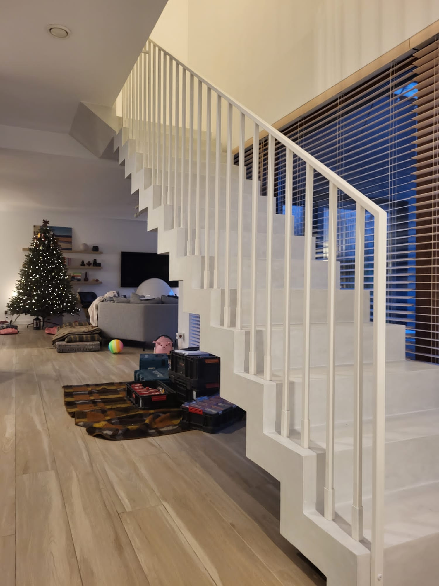 Nowoczesne, białe schody z metalową balustradą w jasnym wnętrzu domu. Widoczne narzędzia sugerują etap montażu. W tle salon z choinką.
