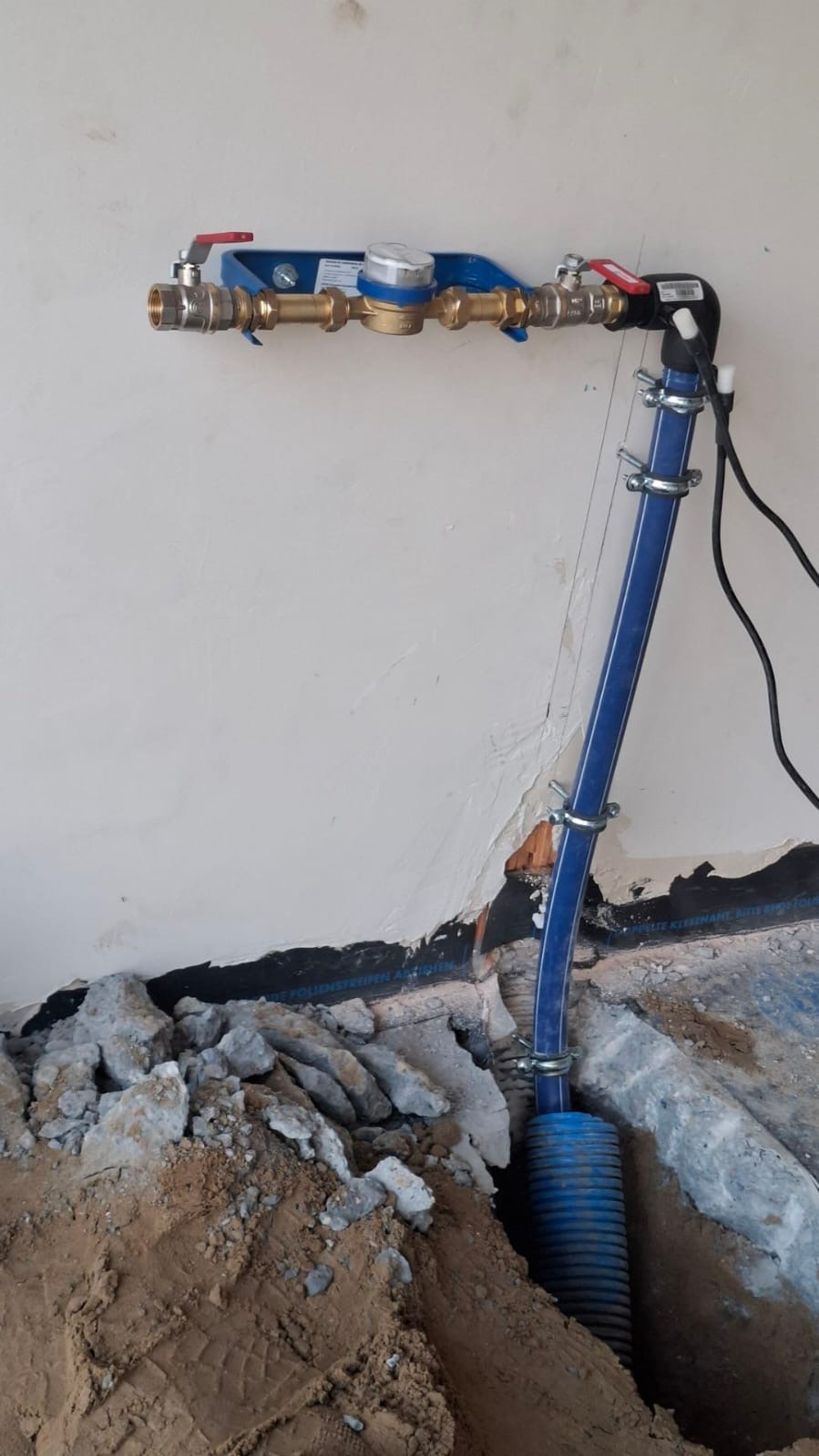 Niebieska rura wodociągowa z licznikiem, zamocowana do ściany, w wykopie ziemnym. Widoczne elementy instalacji i przyłącza. Prace instalacyjne w trakcie realizacji.