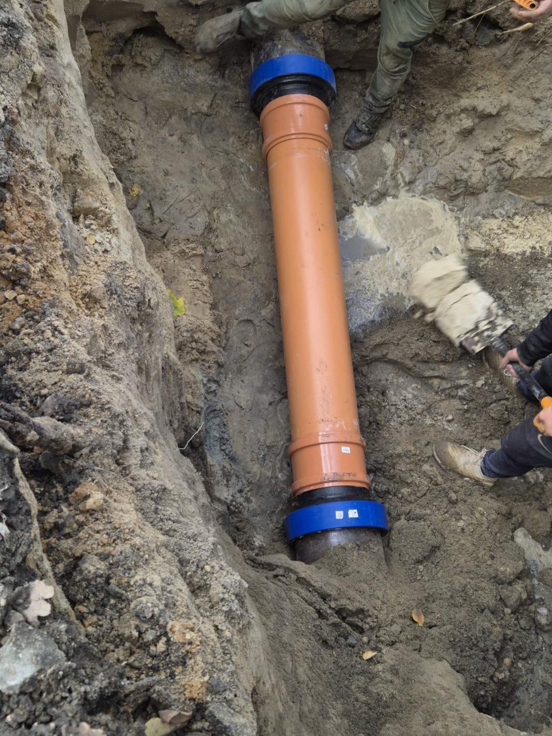 Pomarańczowa rura kanalizacyjna z niebieskimi łącznikami, umieszczona w wykopie ziemnym. Widoczne fragmenty nóg osób wykonujących prace instalacyjne.