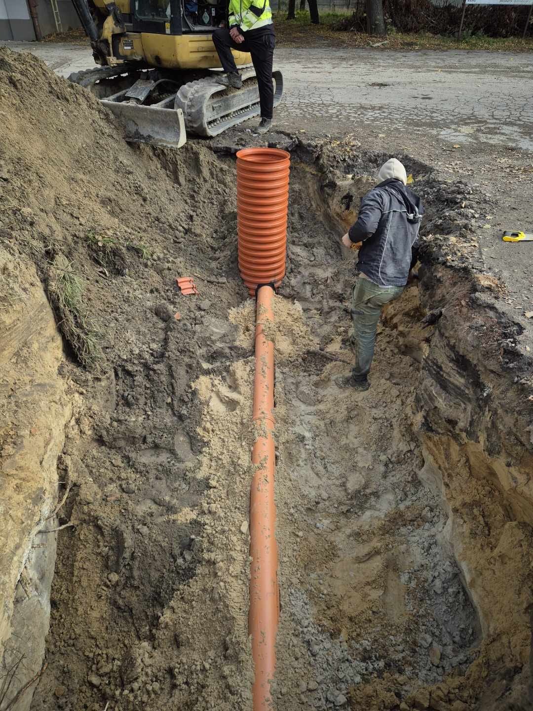 Pomarańczowe rury kanalizacyjne w wykopie ziemnym, widoczne elementy koparki i pracownicy. Instalacja przyłącza wodno-kanalizacyjnego w trakcie realizacji.