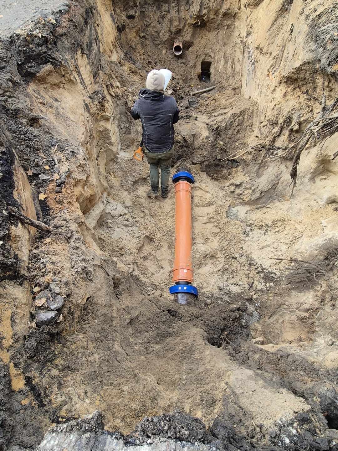 Montaż rury kanalizacyjnej w wykopie ziemnym. Widoczny pracownik z dokumentacją i pomarańczowa rura z niebieskimi łącznikami. Ujęcie z góry, podkreślające głębokość wykopu.