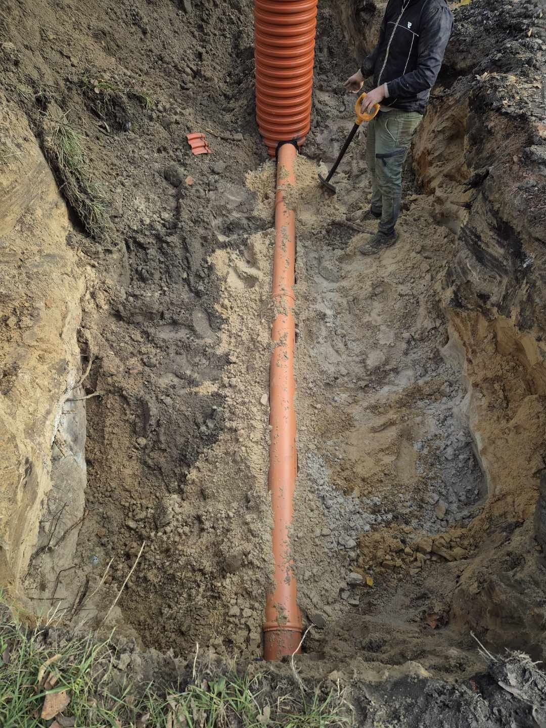 Pomarańczowa rura kanalizacyjna w wykopie ziemnym, połączona z rurą karbowaną. Widoczny fragment osoby z łopatą. Prace ziemne w trakcie realizacji.