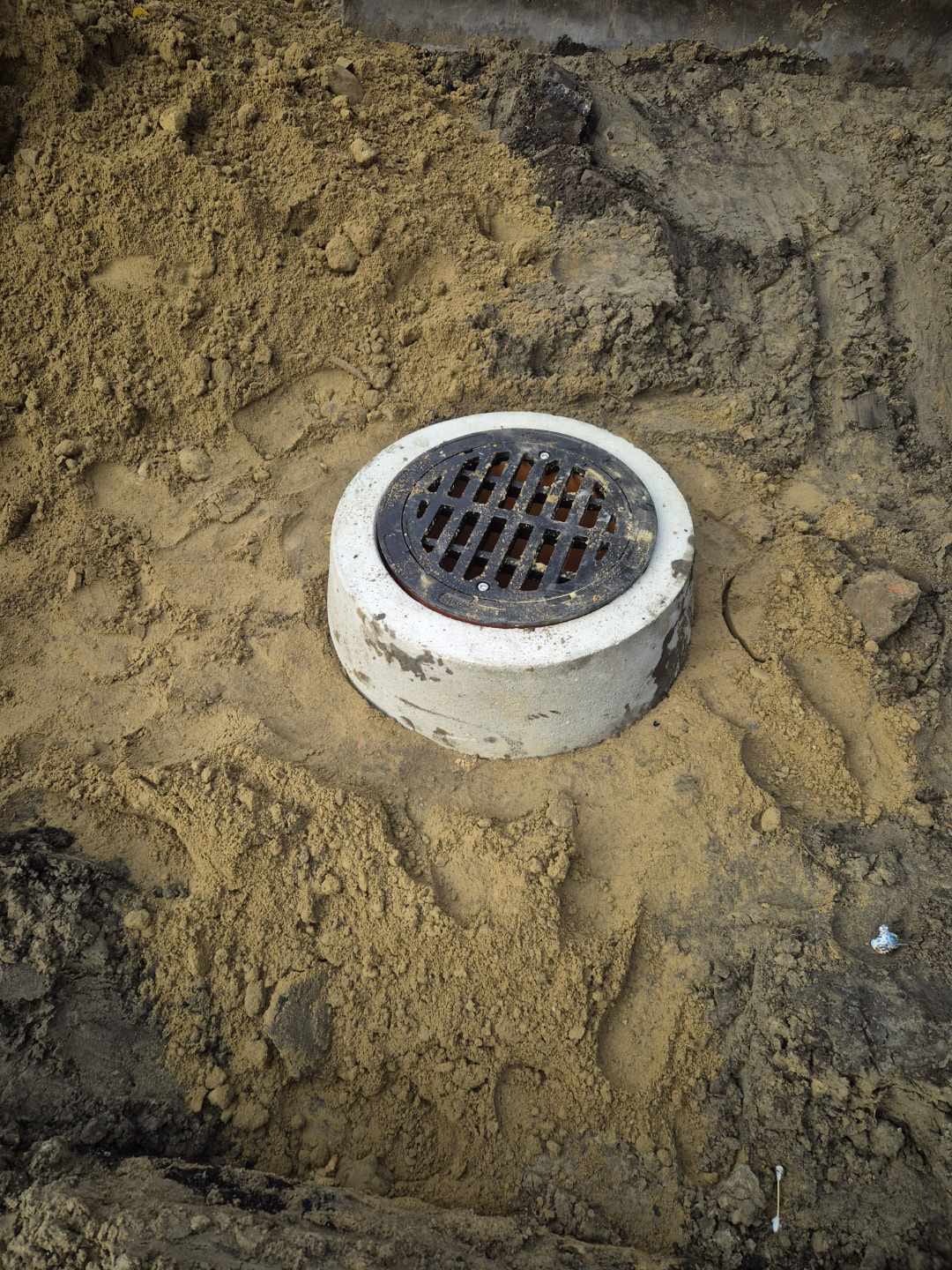 Betonowa studzienka kanalizacyjna z żeliwnym włazem, osadzona w piasku. Widok z góry, ukazujący etap prac instalacyjnych.