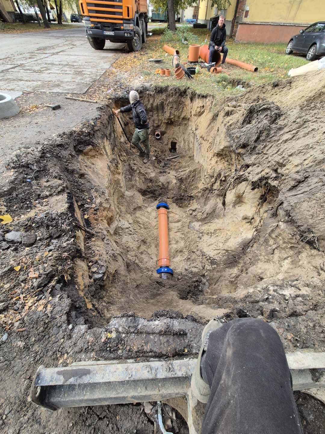 Głęboki wykop w ziemi z widocznymi rurami kanalizacyjnymi, pracownik w środku, pomarańczowa ciężarówka w tle. Prace ziemne i instalacyjne w toku.