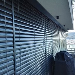 Masters - Rolety - Zamknięte żaluzje fasadowe w kolorze grafitowym na tle nowoczesnego domu z tarasem i widokiem na jezioro. Na tarasie meble ogrodowe i pokrowiec na grilla.