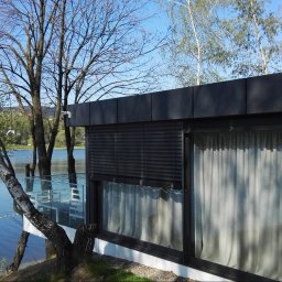 Masters - Rolety - Nowoczesny budynek z dużymi oknami, na których zamontowano poziome żaluzje w kolorze antracytowym, z widokiem na jezioro i drzewa.