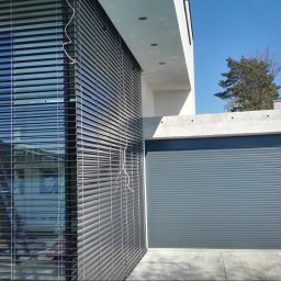 Masters - Rolety - Nowoczesna fasada budynku z szarymi żaluzjami fasadowymi i bramą garażową w tym samym kolorze. Widoczne elementy architektury i zieleń.