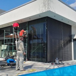 Masters - Rolety - Montaż żaluzji fasadowych na nowoczesnym domu z basenem. Dwóch monterów na rusztowaniu, trzeci obserwuje. Widoczne narzędzia i elementy instalacji.