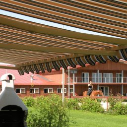 Masters - Rolety - Rozwinięta markiza w paski nad terenem rekreacyjnym w Krakowie. W tle budynek z czerwonym dachem i osoba na koniu.