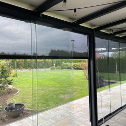 Masters - Rolety - Zadaszenie tarasu z czarną konstrukcją i rozsuwanymi przesłonami w Krakowie. Widok na zadbany ogród przez szklane panele.