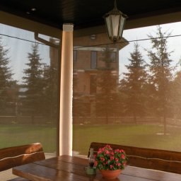 Masters - Rolety - Zadaszony taras z drewnianym stołem i ławkami, osłonięty brązowymi, zwijanymi przesłonami. W tle widoczne drzewa iglaste i budynek.