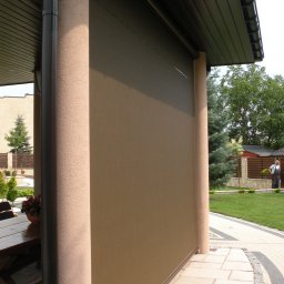 Masters - Rolety - Beżowa, zwinięta osłona przeciwsłoneczna na tarasie, widoczna z boku, z elementami architektury ogrodowej w tle i fragmentem drewnianego stołu.