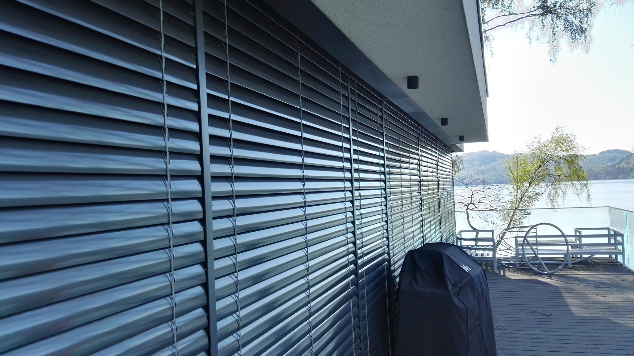 Zamknięte żaluzje fasadowe w kolorze grafitowym na tle nowoczesnego domu z tarasem i widokiem na jezioro. Na tarasie meble ogrodowe i pokrowiec na grilla.