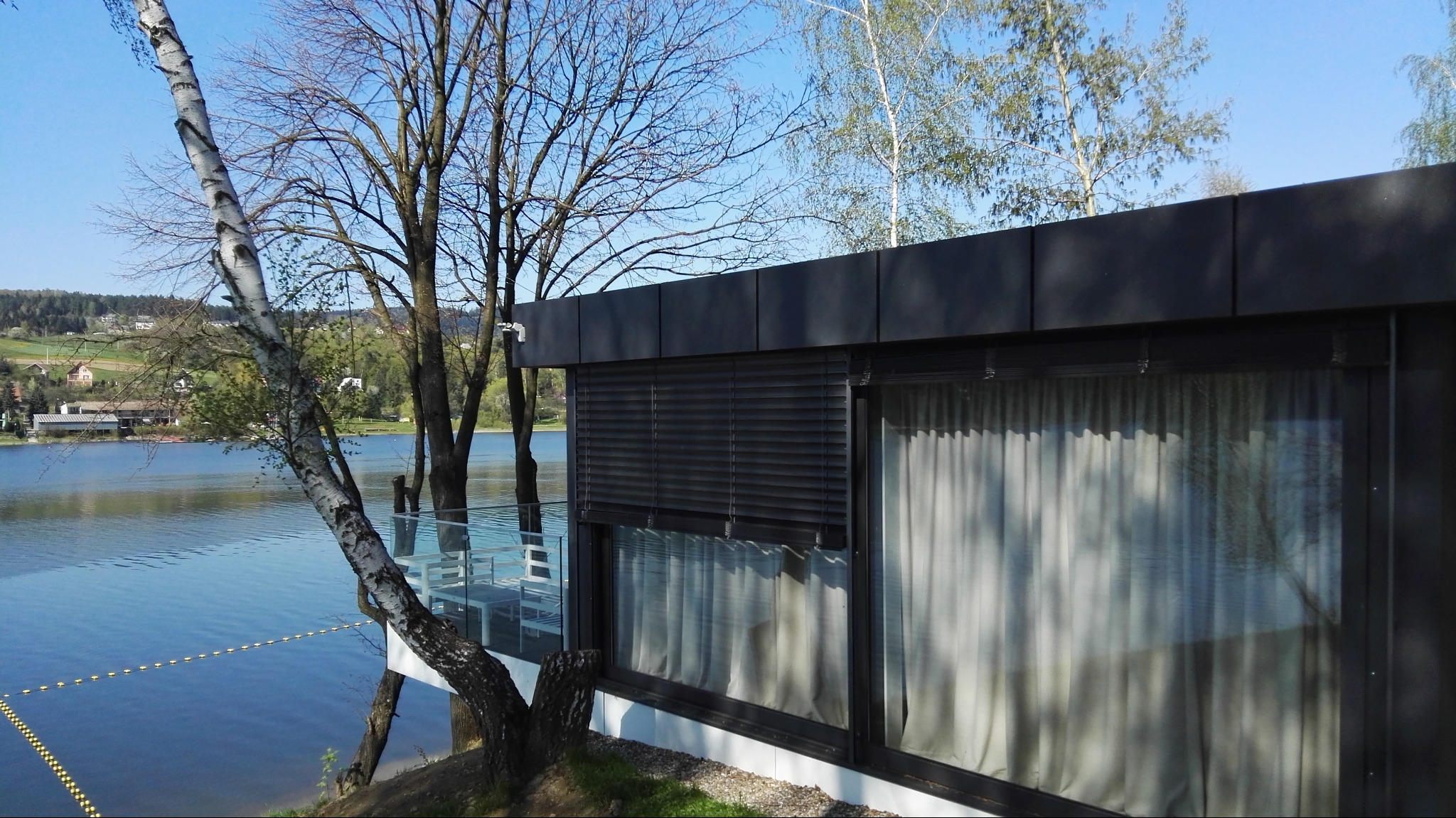 Nowoczesny budynek z dużymi oknami, na których zamontowano poziome żaluzje w kolorze antracytowym, z widokiem na jezioro i drzewa.