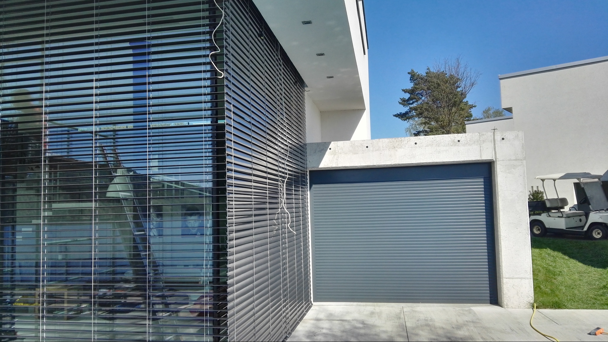 Nowoczesna fasada budynku z szarymi żaluzjami fasadowymi i bramą garażową w tym samym kolorze. Widoczne elementy architektury i zieleń.