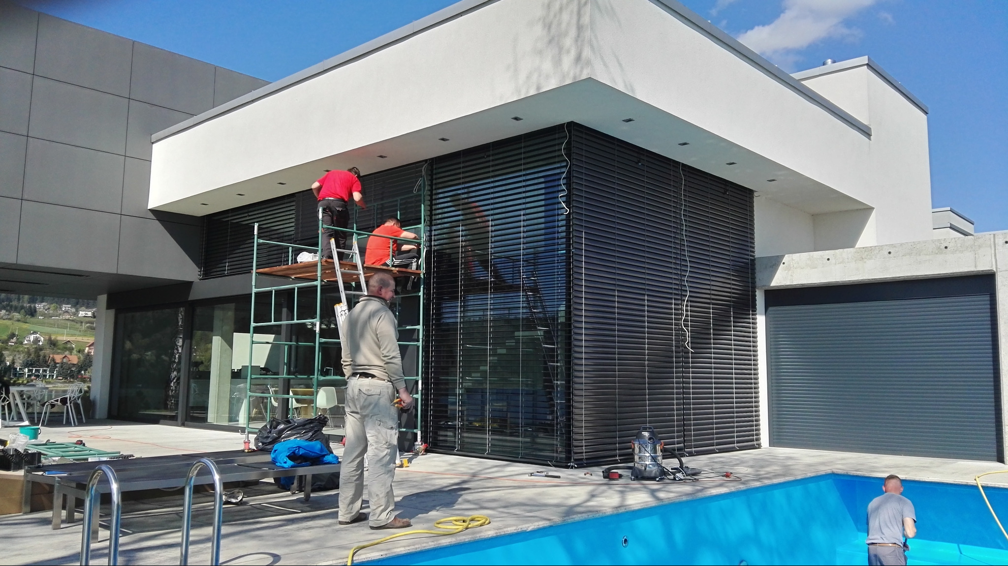 Montaż żaluzji fasadowych na nowoczesnym domu z basenem. Dwóch monterów na rusztowaniu, trzeci obserwuje. Widoczne narzędzia i elementy instalacji.