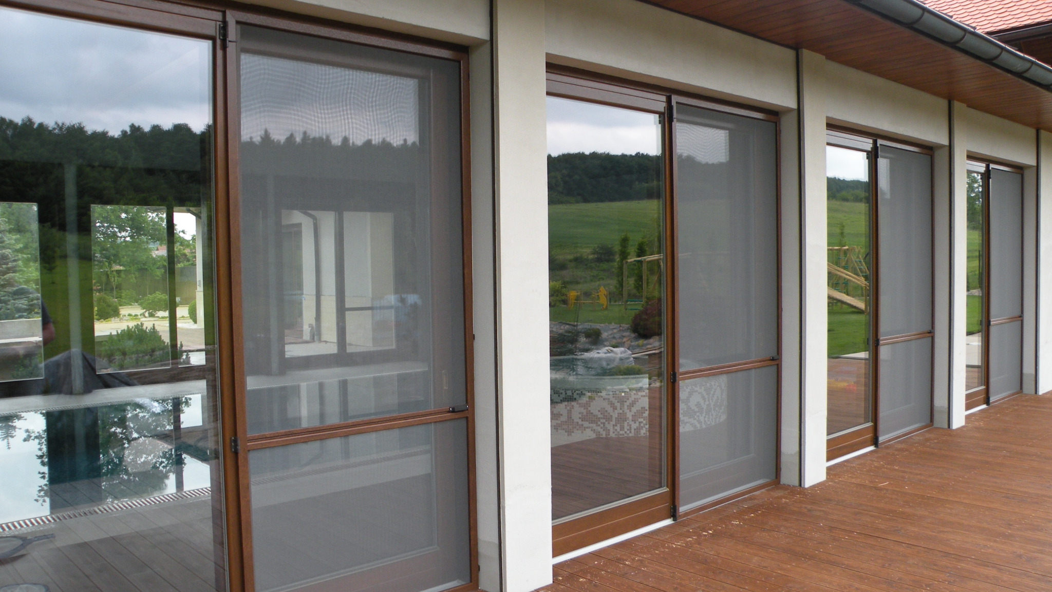 Zamontowane moskitiery ramkowe w oknach tarasowych, brązowe ramy, widok z zewnątrz na taras i ogród. Realizacja w Krakowie.