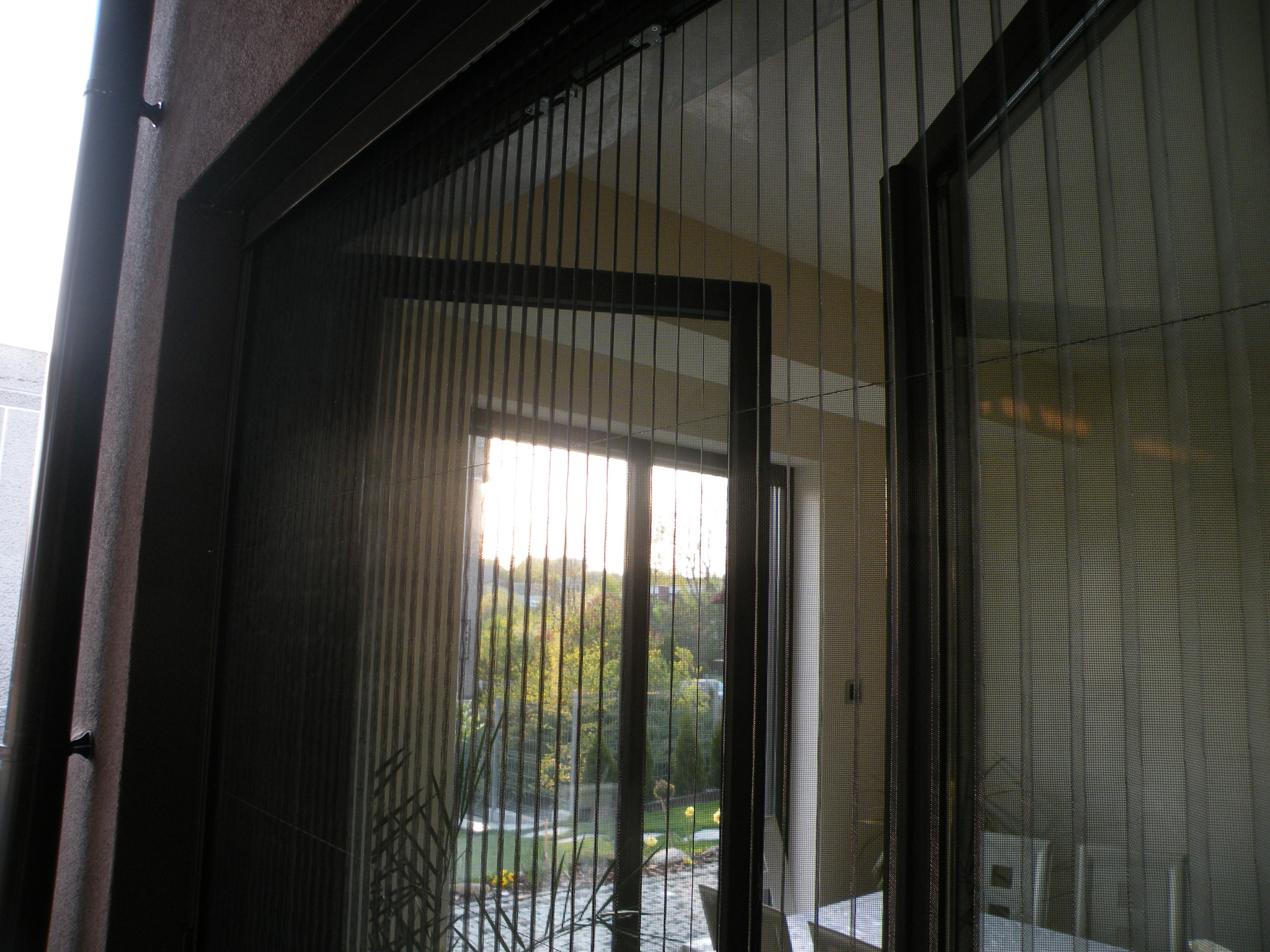 Zamknięta, czarna moskitiera ramkowa na balkonowych drzwiach, z widokiem na ogród przez siatkę. Delikatne światło słoneczne wpada do wnętrza.