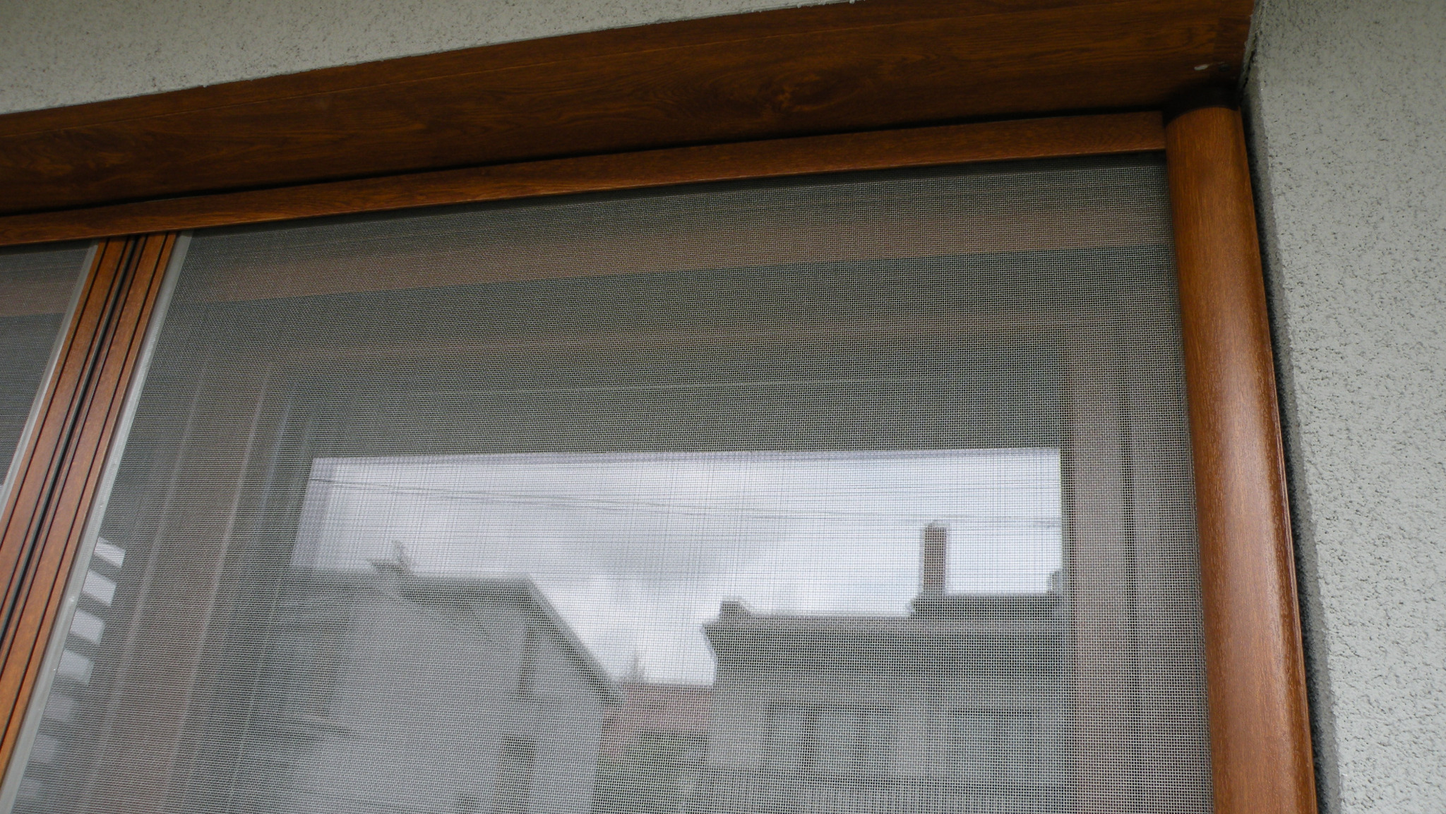 Zamknięta moskitiera okienna z brązową ramą, widok z bliska na siatkę i odbicie otoczenia w oknie. Teksturowana ściana w tle.