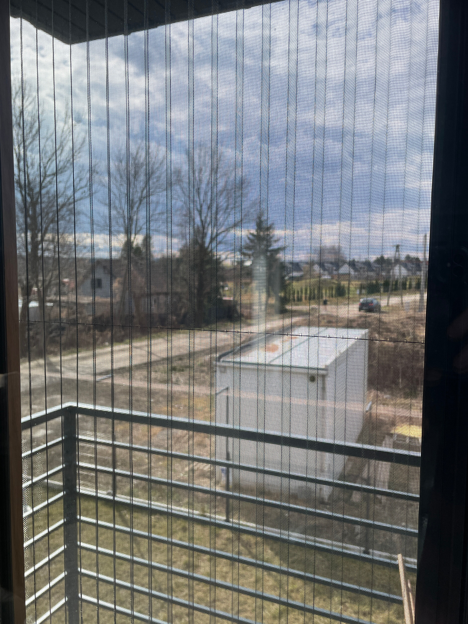 Zamknięta moskitiera plisowana na balkonie, widok na zewnątrz z zabudową i drzewami. Delikatna siatka moskitiery z bliska.