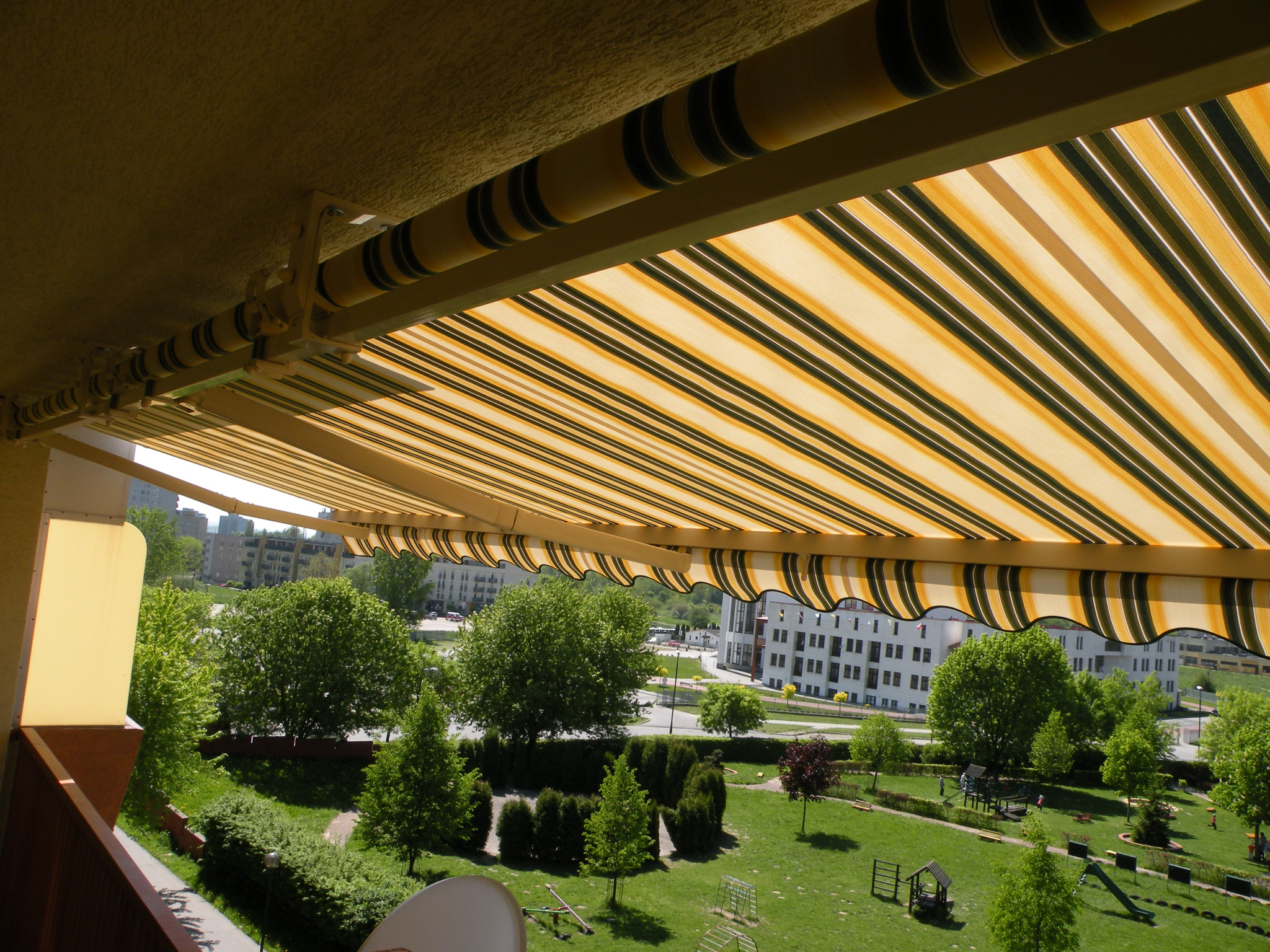 Tarasowa markiza w paski, zamontowana nad balkonem, z widokiem na zielony park miejski i budynki w tle, w słoneczny dzień.