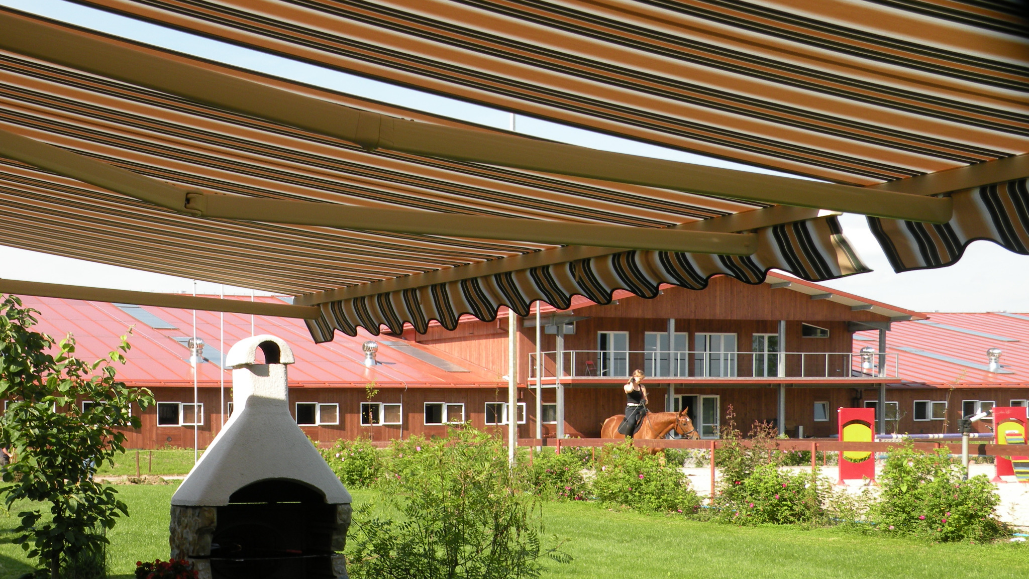 Rozwinięta markiza w paski nad terenem rekreacyjnym w Krakowie. W tle budynek z czerwonym dachem i osoba na koniu.