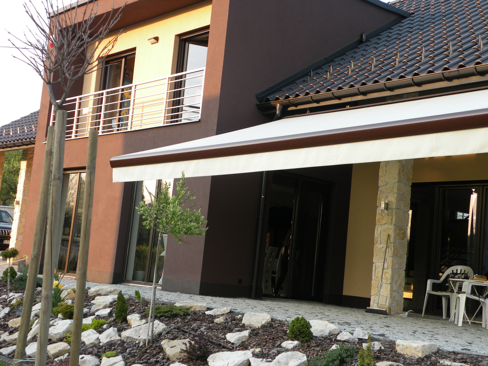 Elegancki dom z beżową markizą nad wejściem, widok z ogrodu. Jasne płytki chodnikowe i starannie zaaranżowana zieleń podkreślają nowoczesny charakter budynku.