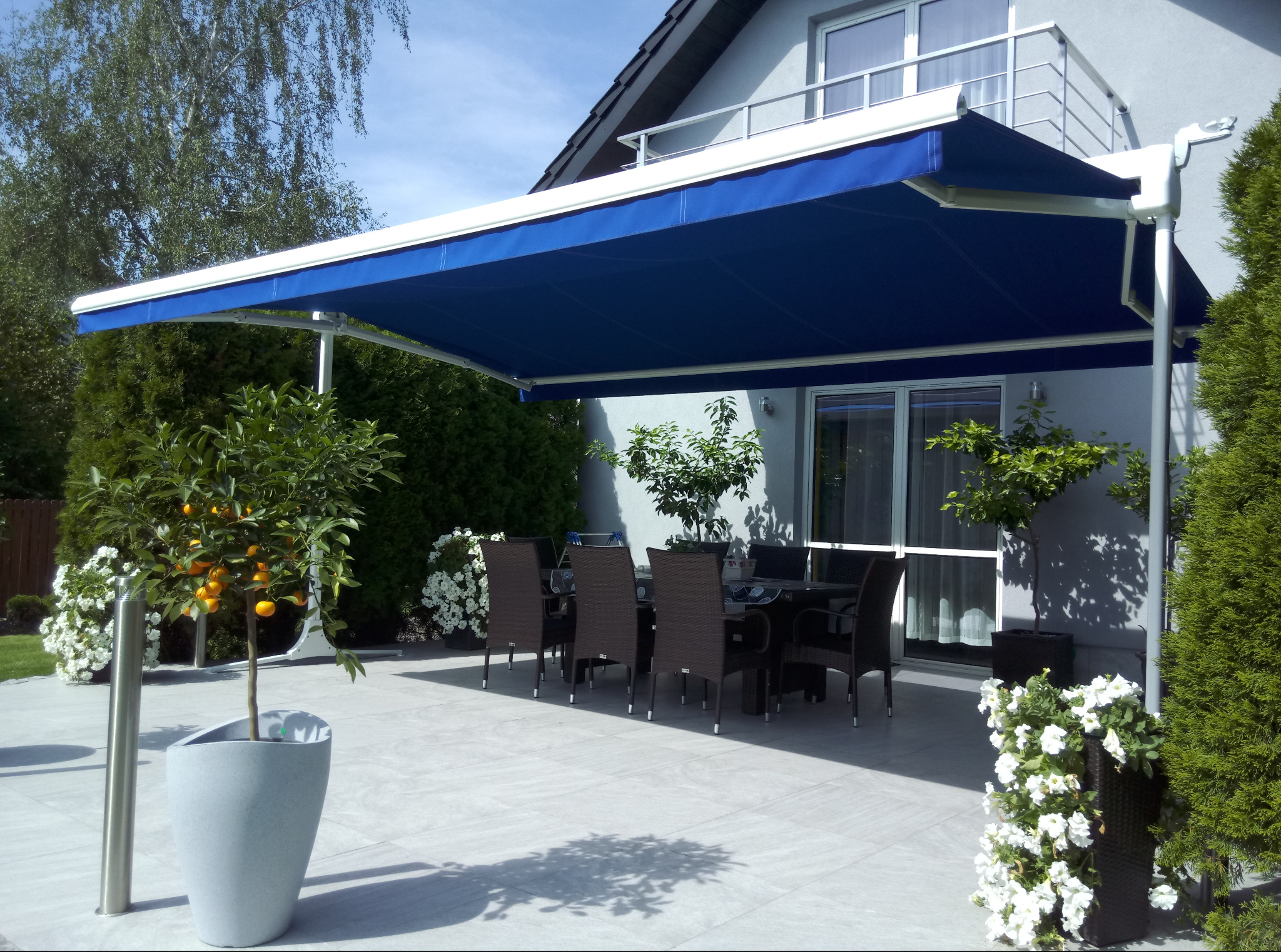 Błękitna markiza tarasowa z białym stelażem, osłaniająca stół z krzesłami na jasnym patio. W tle dom z balkonem i zadbana zieleń.