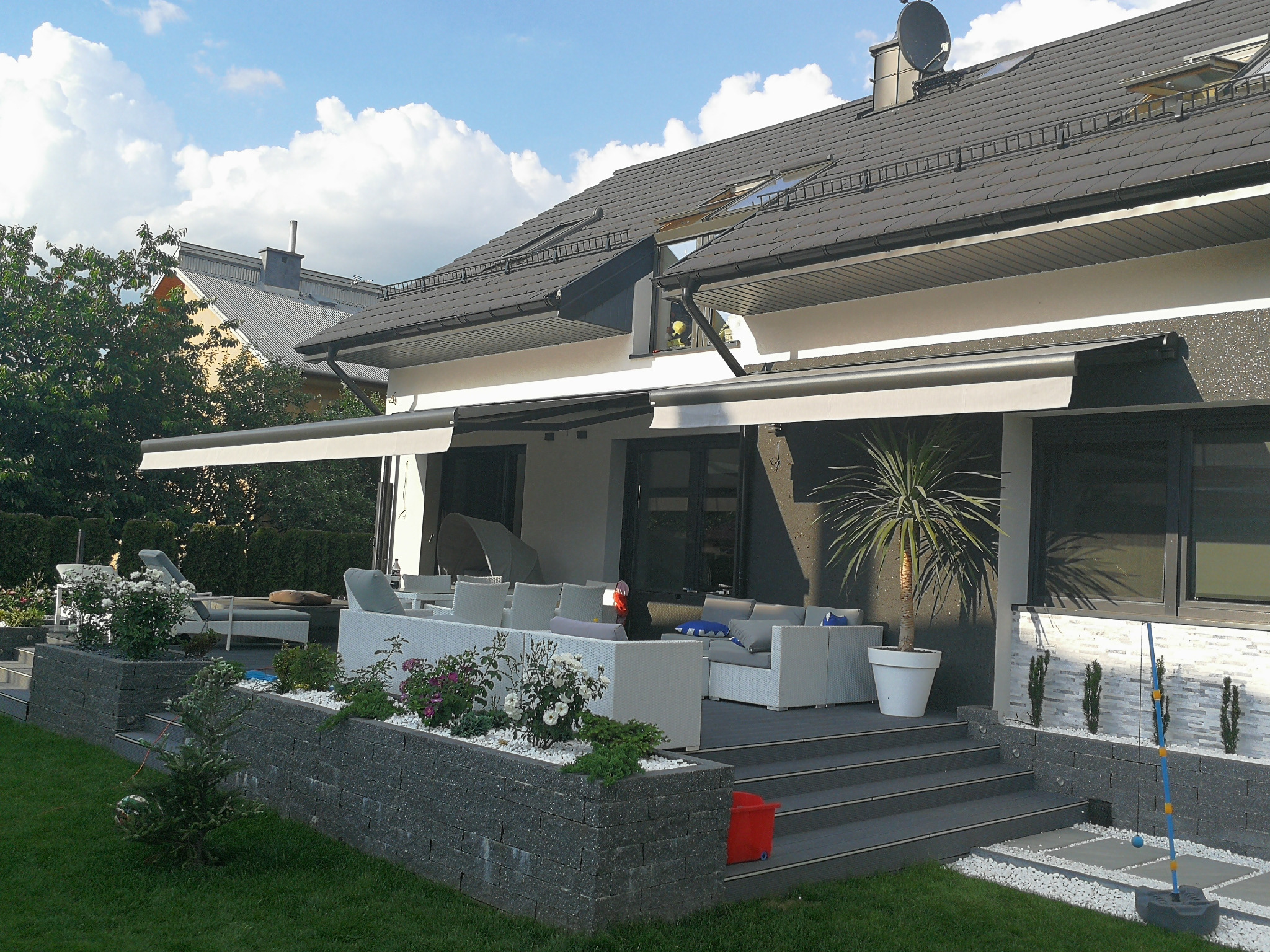 Elegancki taras z markizami w nowoczesnym domu. Białe meble ogrodowe, rośliny w donicach i kamienne obrzeża tworzą spójną całość. Widok na błękitne niebo z chmurami.