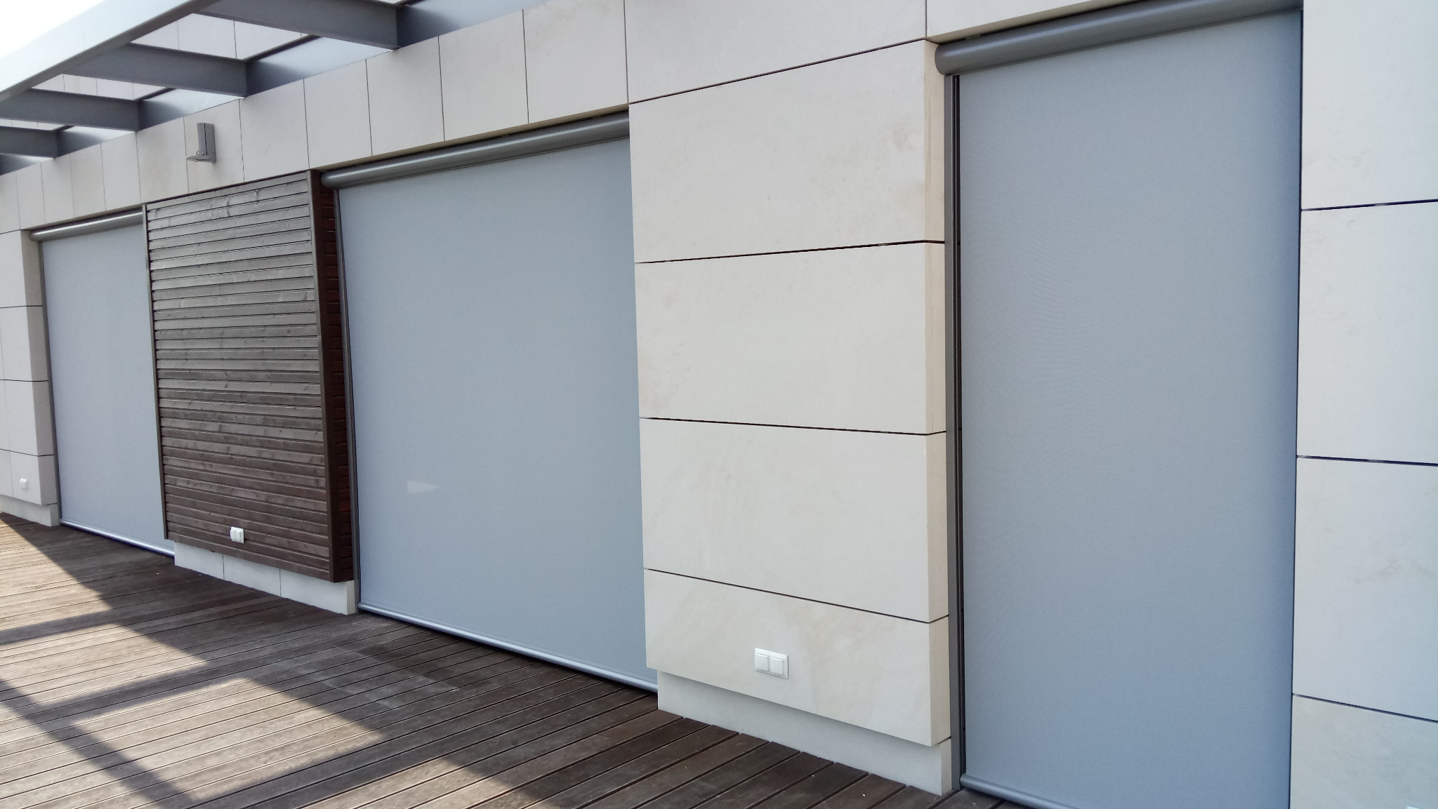 Szare, zwinięte przesłony okienne na tarasie z drewnianą podłogą. Widoczne elementy elewacji z jasnego kamienia i drewniane panele. Stonowana kolorystyka.