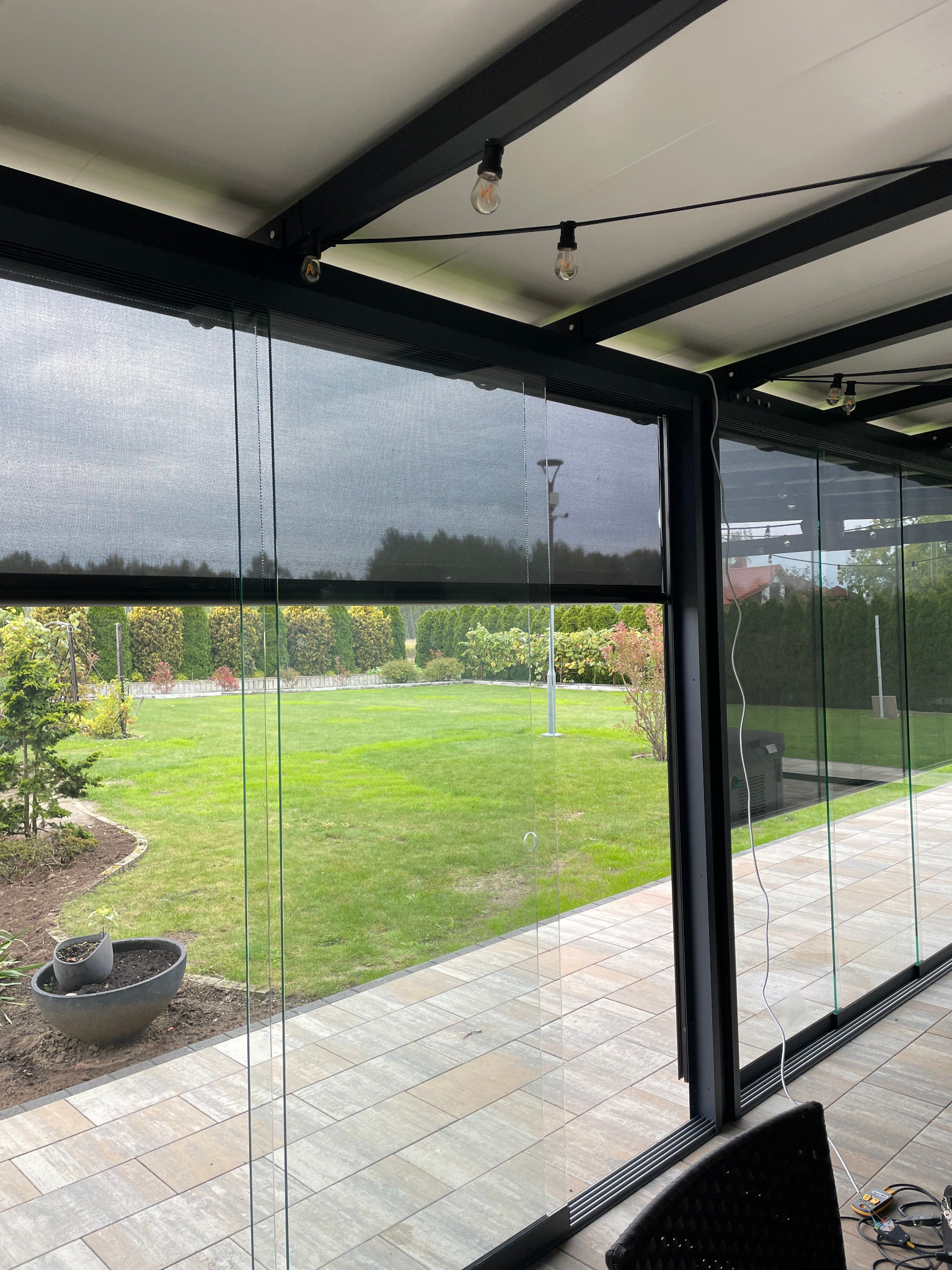 Zadaszenie tarasu z czarną konstrukcją i rozsuwanymi przesłonami w Krakowie. Widok na zadbany ogród przez szklane panele.