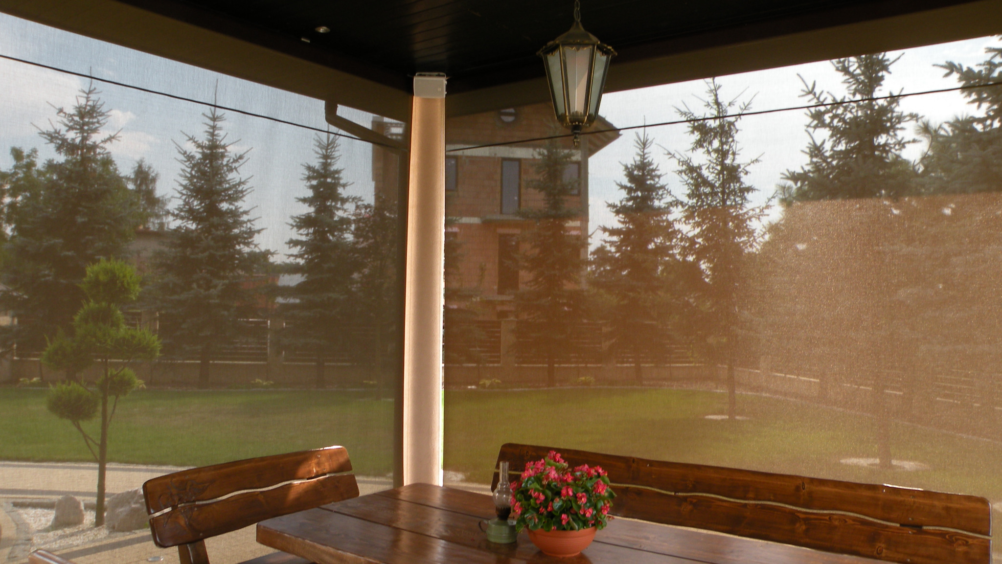 Zadaszony taras z drewnianym stołem i ławkami, osłonięty brązowymi, zwijanymi przesłonami. W tle widoczne drzewa iglaste i budynek.