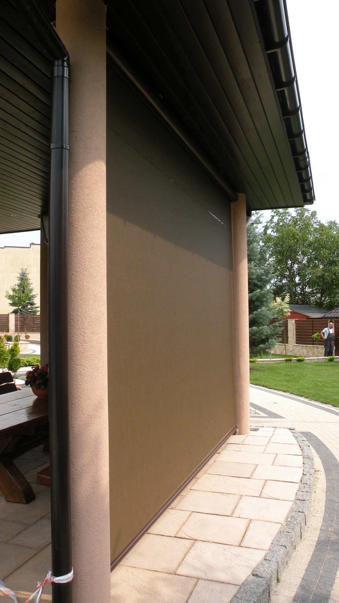 Beżowa, zwinięta osłona przeciwsłoneczna na tarasie, widoczna z boku, z elementami architektury ogrodowej w tle i fragmentem drewnianego stołu.