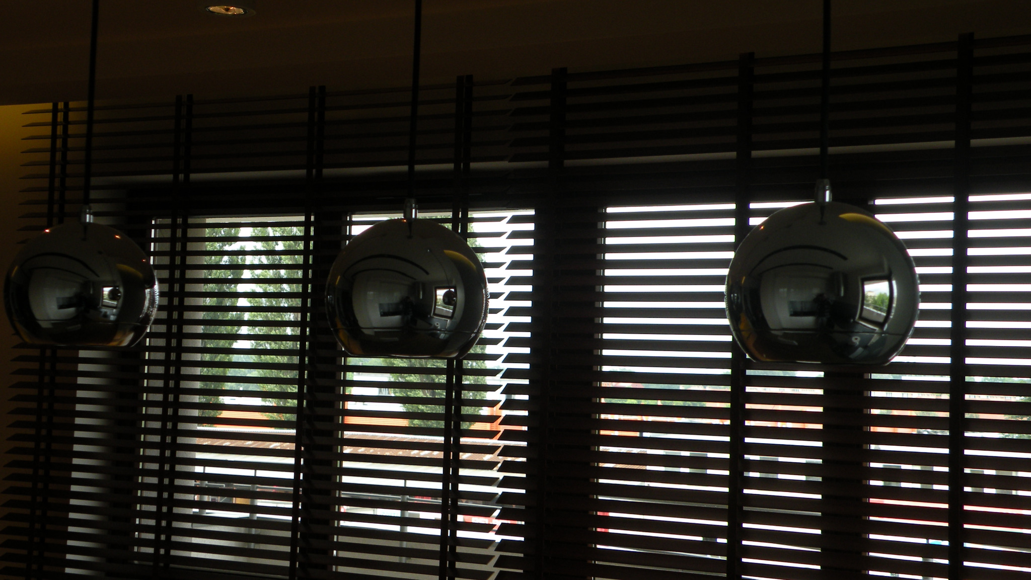 Drewniane żaluzje poziome w oknie, z widokiem na zieleń, oświetlone przez trzy metalowe, kuliste lampy wiszące. Ciemne drewno lameli tworzy regularny wzór.